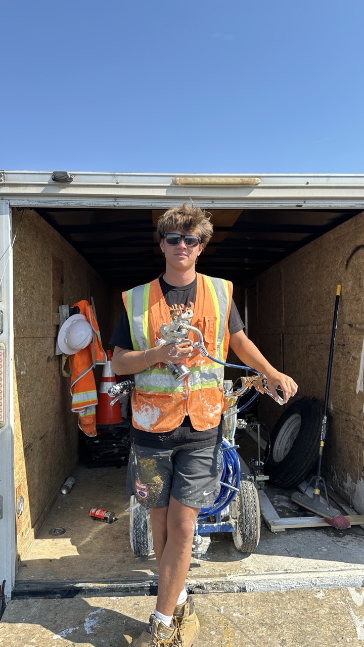 Caden in a safety vest standing beside a Premier Striping equipment trailer